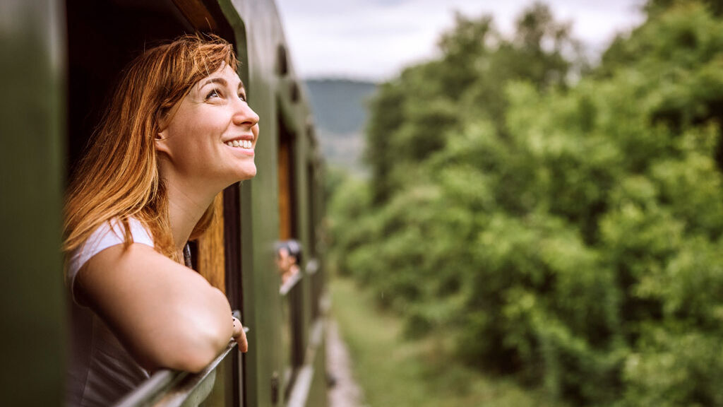 Eine Frau schaut aus einem Zug-Fenster nach draußen in die Natur und freut sich.