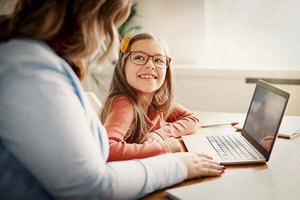 Ein junges Mädchen, welches eine Brille trägt, sitzt mit ihrer Mutter vor einem Laptop. Sie lächeln sich an. 
