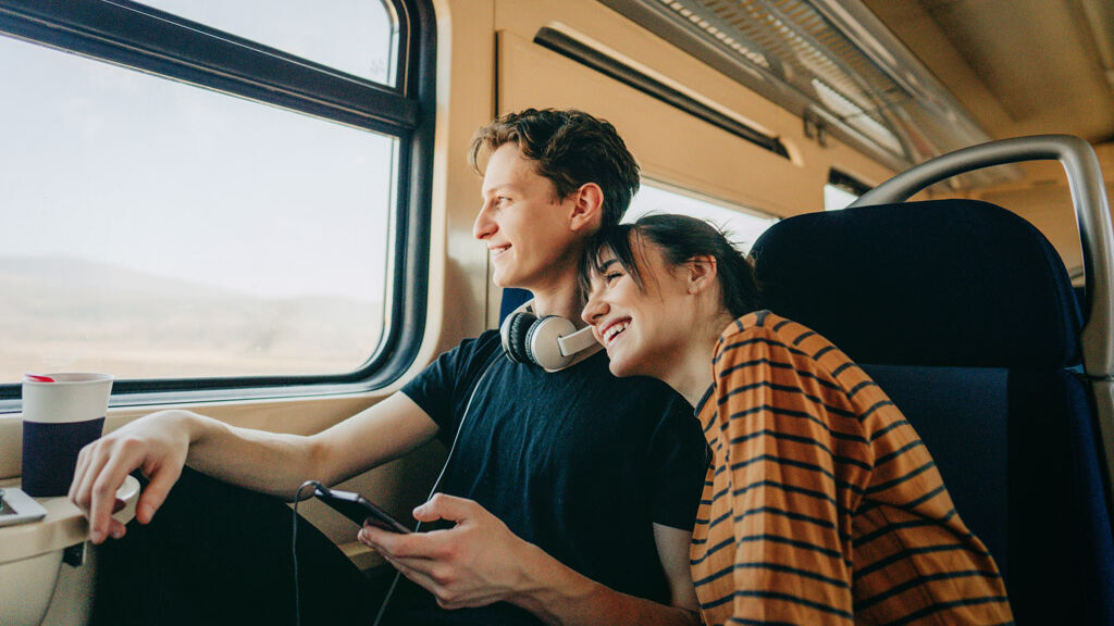 ein junges Paar sitzt gemeinsam im Zug und sie schauen fröhlich aus dem Fenster.