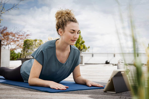 Eine Frau macht auf einer Dachterasse Yoga-Übungen über einen digitalen Gesundheitskurs auf ihrem Tablet. 