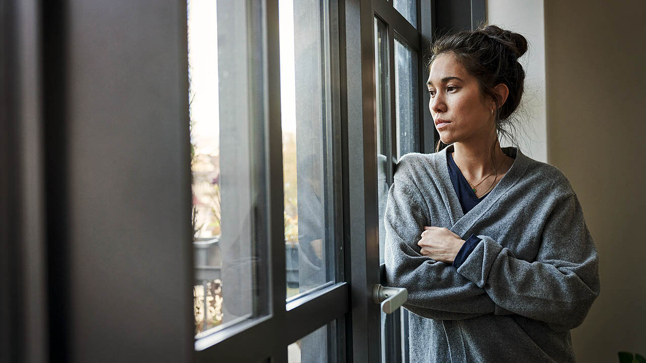 Eine Frau steht mit verschränkten Armen ans Fenster gelehnt und sieht nachdenklich und traurig nach draußen.