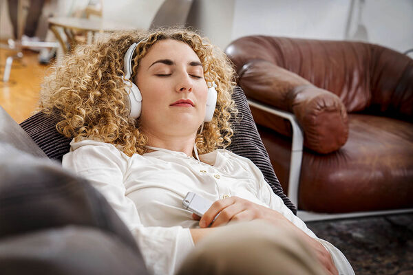 Eine Frau liegt mit Kopfhörer auf dem Sofa, hält ihr Handy in der Hand und entspannt mit geschlossenen Augen.