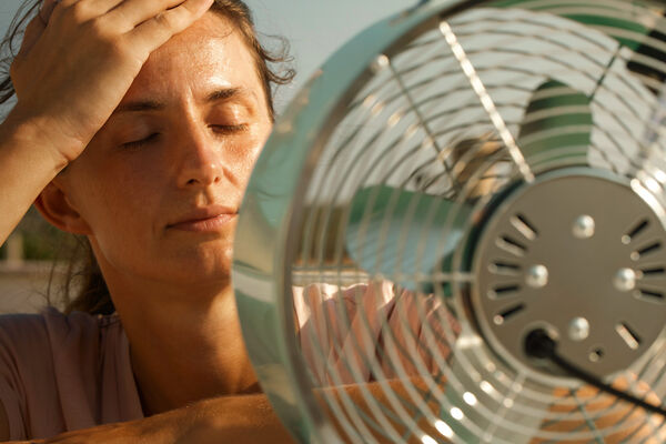 Schwitzende junge Frau vor Ventilator fasst sich an den Kopf