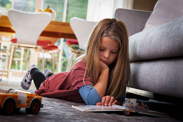 Ein Mädchen liegt auf dem Bauch auf dem Boden im Wohnzimmer und liest ein Buch.