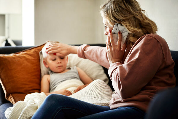 Eine Mutter sitzt mit ihrem kranken Sohn auf dem Sofa und misst mit ihrer Handfläche, ob er Fieber hat. Sie telefoniert währenddessen.