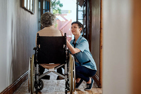 Eine Pflegefachkraft sitzt in der Hocke neben einer älteren Frau im Rollstuhl und berührt sie an der Schulter.