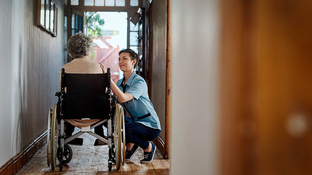 Eine Pflegefachkraft sitzt in der Hocke neben einer älteren Frau im Rollstuhl und berührt sie an der Schulter.