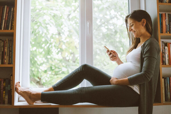 Eine schwangere Frau sitzt mit ihrem Mobiltelefon auf der Fensterbank und lächelt.