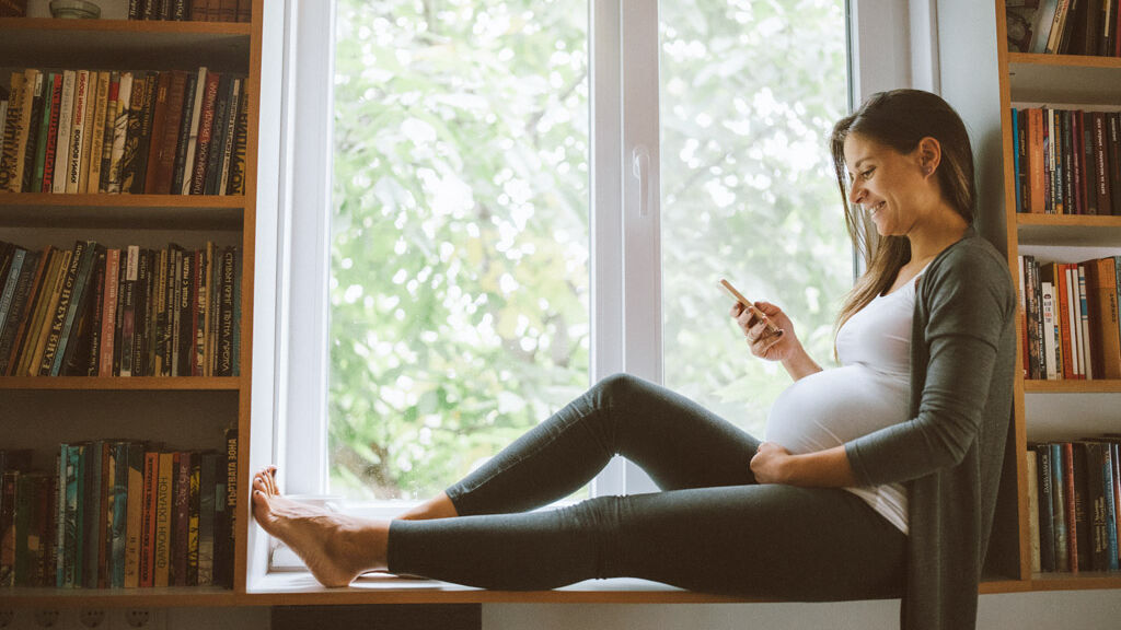 Eine schwangere Frau sitzt mit ihrem Mobiltelefon auf der Fensterbank und lächelt.