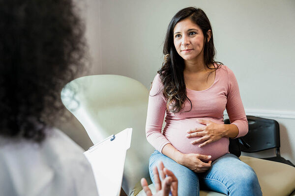 Eine schwangere Frau sitzt auf einer Behandlungsliege einer Arztpraxis und wird von einer Ärztin beraten.