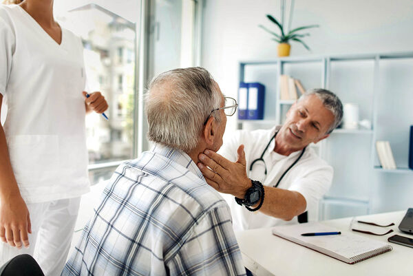 Ein Arzt untersucht in seinem Behandlungszimmer einen älteren Mann durch eine Tastuntersuchung am Hals.