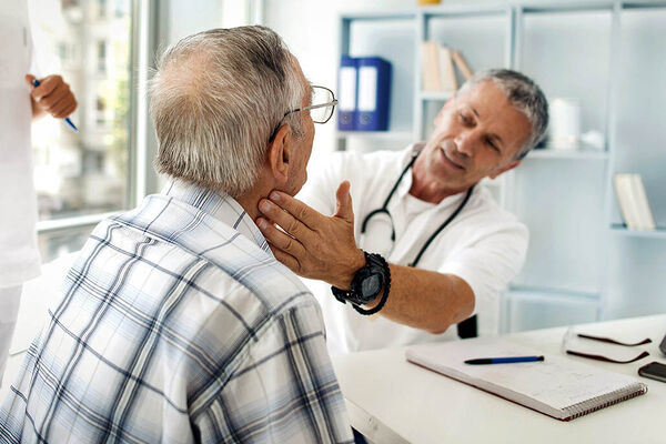 Ein Arzt untersucht in seinem Behandlungszimmer einen älteren Mann durch eine Tastuntersuchung am Hals.