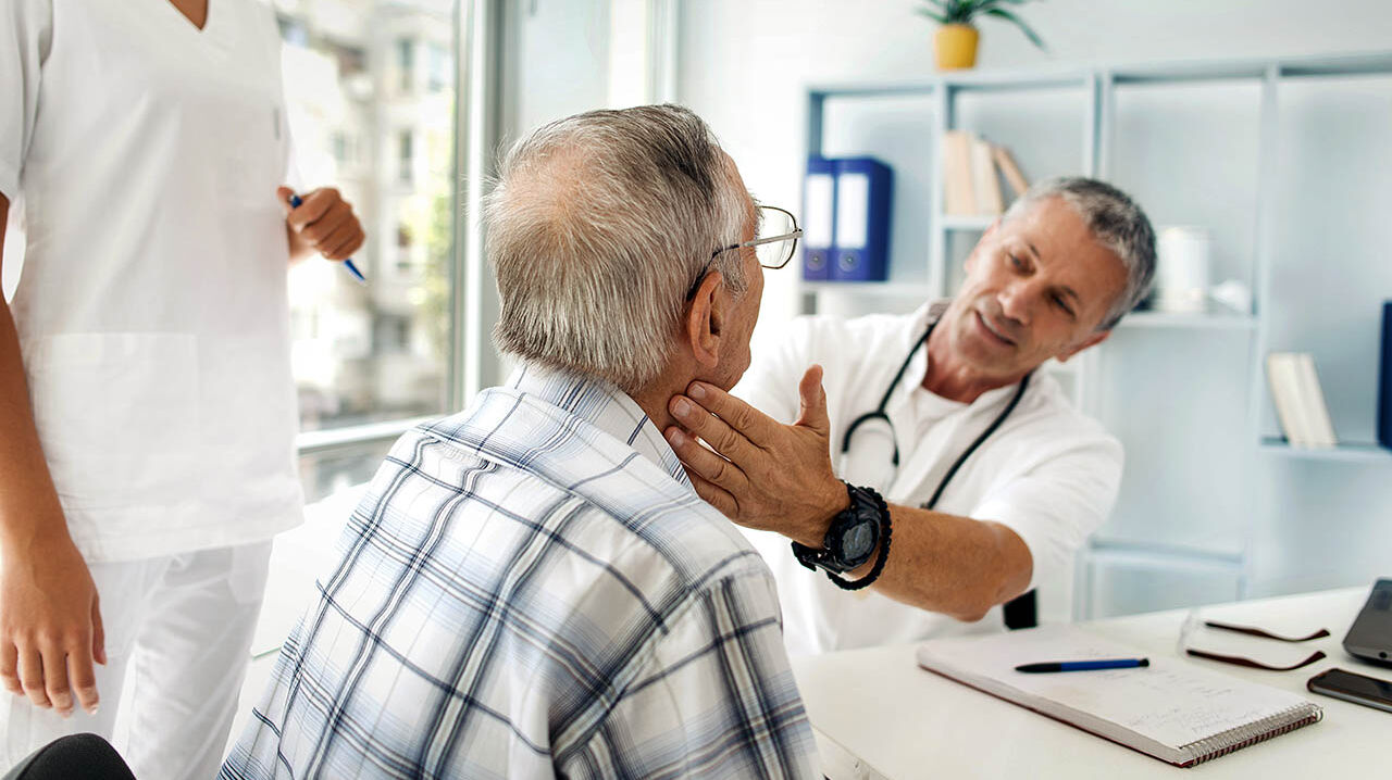 Ein Arzt untersucht in seinem Behandlungszimmer einen älteren Mann durch eine Tastuntersuchung am Hals.