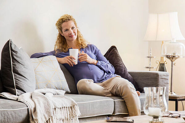 Eine schwangere Frau sitzt auf dem Sofa im Wohnzimmer und trinkt eine Tasse Tee.