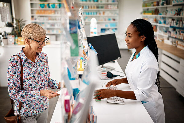 Eine Frau löst ihr Arzneimittelrezept in einer Apotheke ein und wird von einer Apothekerin über den Tresen beraten.