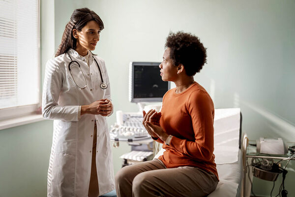 Eine Patientin sitzt auf einer Liege im Behandlungsraum und erklärt ihre Symptome der Ärztin.