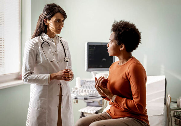 Eine Patientin sitzt auf einer Liege im Behandlungsraum und erklärt ihre Symptome der Ärztin.