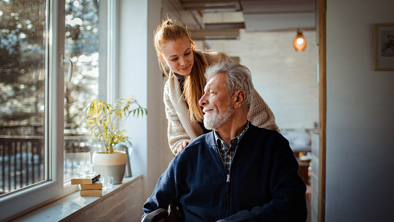 Ein älterer Mann sitzt im Rollstuhl zu Hause und wird von seiner Tochter geschoben.