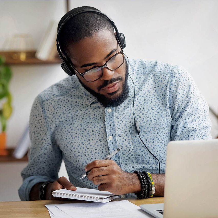 Ein Mann sitzt mit Kopfhörer und Notizblock vor seinem Laptop und hört interessiert zu.