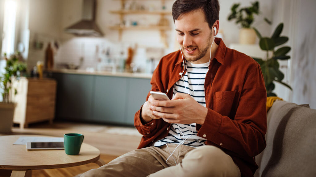 Ein Mann sitzt auf dem Sofa und hört etwas über Kopfhörer und sein Mobiltelefon an.