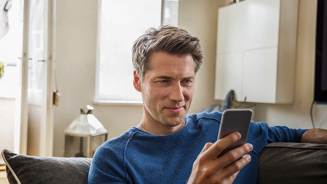 Ein Mann sitzt im Wohnzimmer auf dem Sofa und schaut in sein Handy.