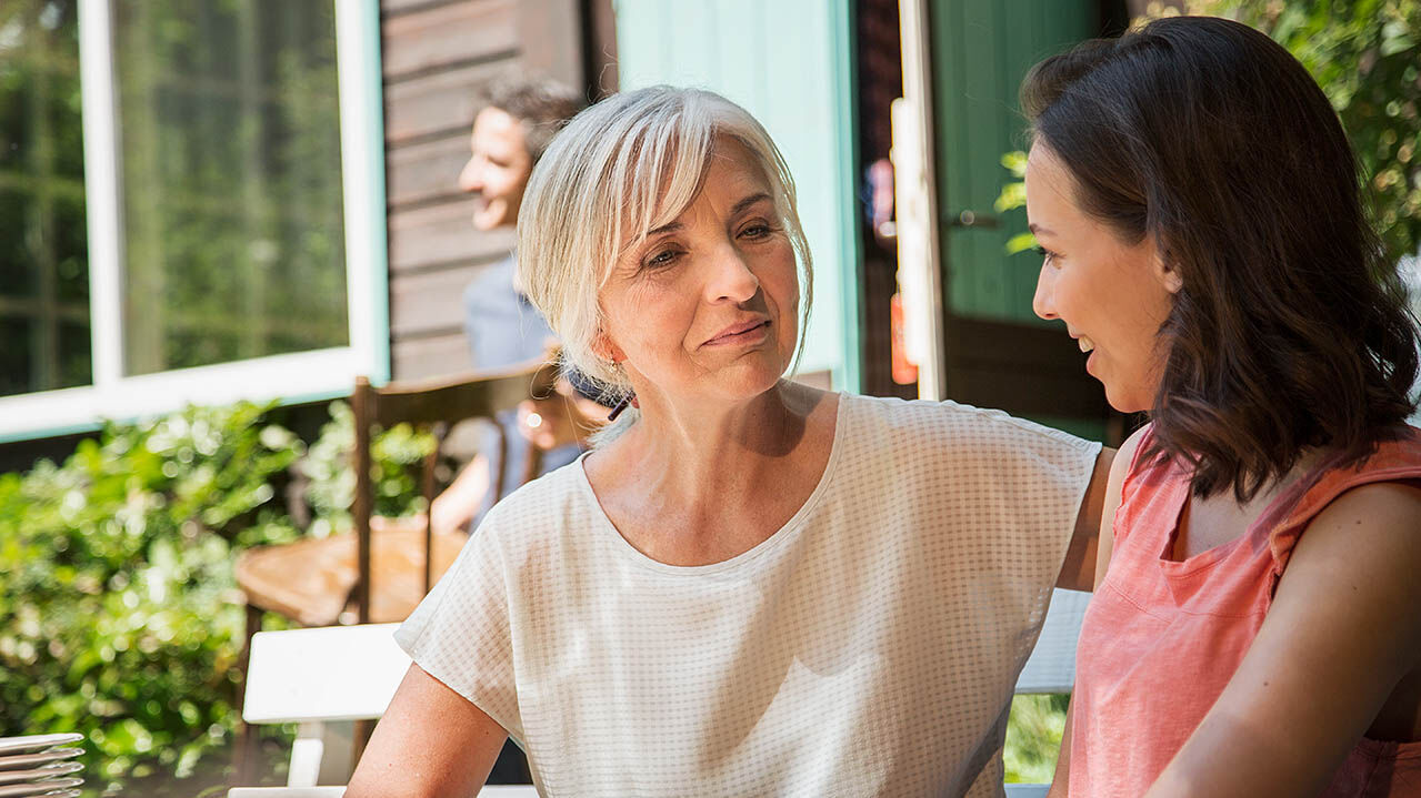 Eine ältere Frau sitzt mit ihrer Tochter auf einer Bank und sie unterhalten sich.