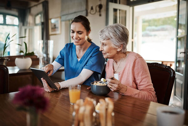 Eine Pflegefachkraft sitzt mit einer älteren Frau am Esstisch und zeigt ihr etwas auf dem Tablet.