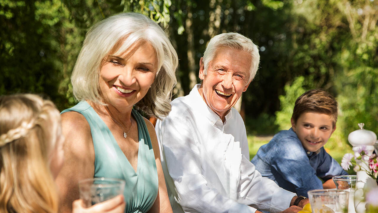 Großeltern sitzen mit ihren Enkeln am Gartentisch und lachen gemeinsam.
