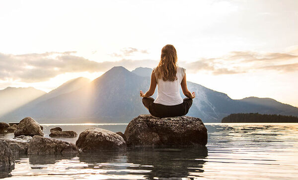 Eine junge Frau sitzt auf einem Stein an einem See und meditiert.