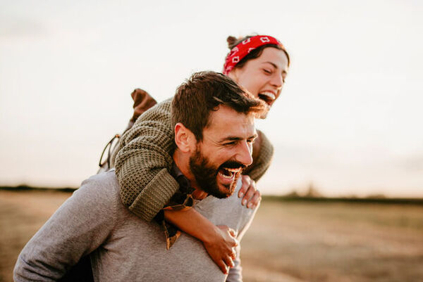Ein Mann trägt eine Frau im Huckepack auf dem Rücken. Die beiden sind in der Natur und lachen herzlich.