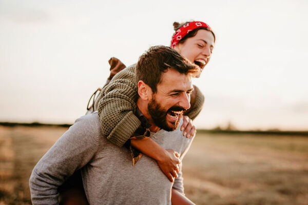 Ein Mann trägt eine Frau im Huckepack auf dem Rücken. Die beiden sind in der Natur und lachen herzlich.