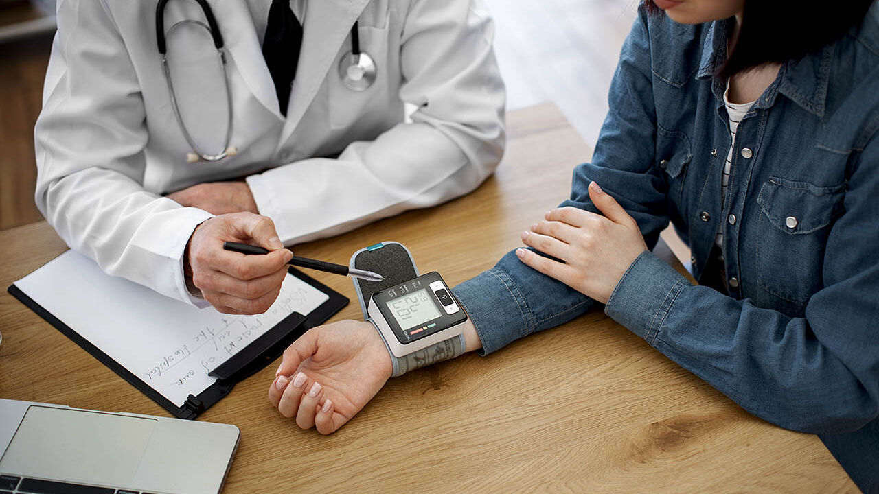 Ein Arzt und eine Patientin sitzen gemeinsam an einem Tisch und messen mit einem Gerät an ihrem Handgelenk ihren Blutdruck.
