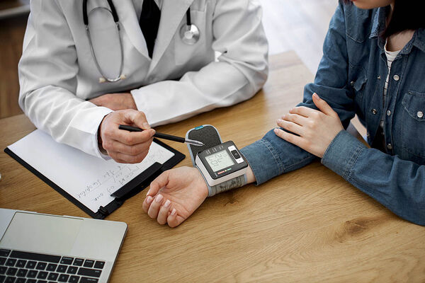 Ein Arzt und eine Patientin sitzen gemeinsam an einem Tisch und messen mit einem Gerät an ihrem Handgelenk ihren Blutdruck.