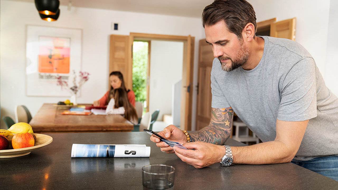 Ein Mann lehnt am Küchentresen und gibt seine Versichertennummer von seiner elektronischen Gesundheitskarte in sein Handy ein.