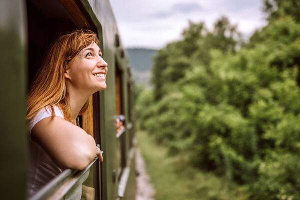 Eine Frau schaut aus einem Zug-Fenster nach draußen in die Natur und freut sich.