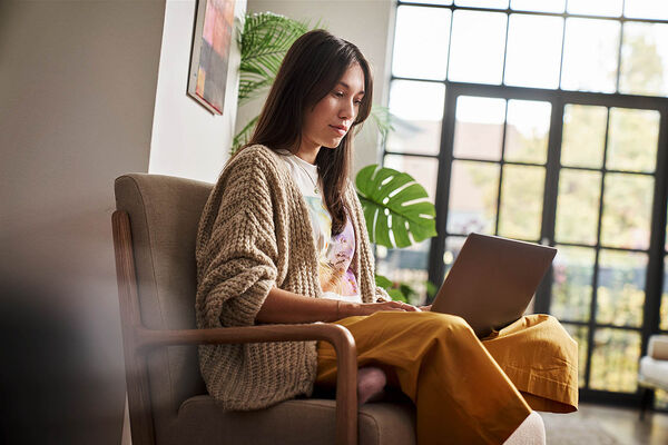 Eine Frau sitzt auf einem Sessel im Wohnzimmer und arbeitet an ihrem Laptop.