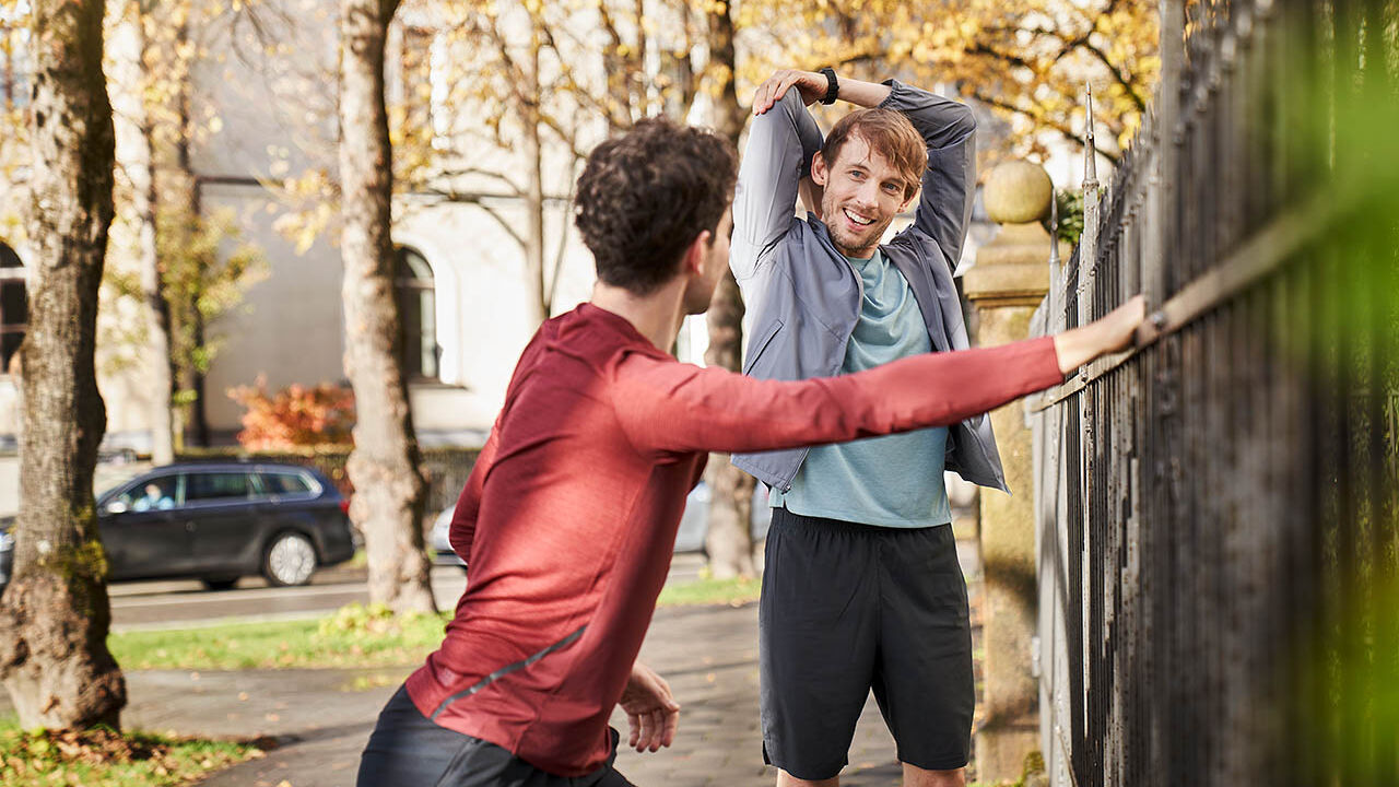 Zwei Männer dehnen sich, bevor sie an einem sonnigen, herbstlichen Tag gemeinsam joggen gehen.