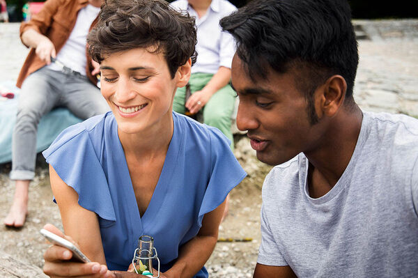 Eine Frau sitzt draußen an einem Fluss und zeigt einem Mann etwas auf ihrem Handy.