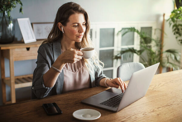 Eine Frau sitzt mit Kopfhörer und einer Tasse Tee vor ihrem Laptop im Esszimmer.