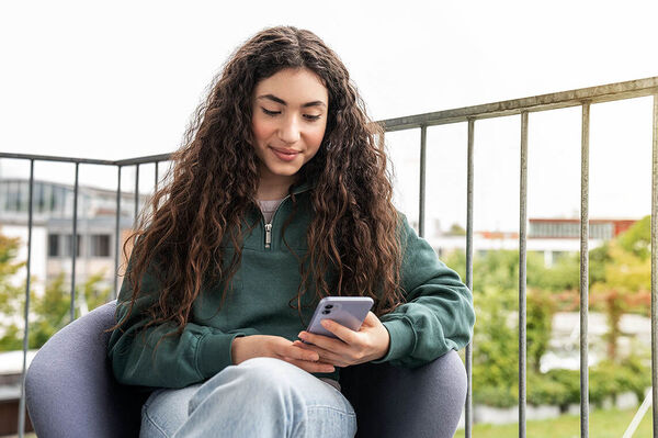 Ein Mädchen sitzt auf dem Balkon und schaut in ihr Handy.
