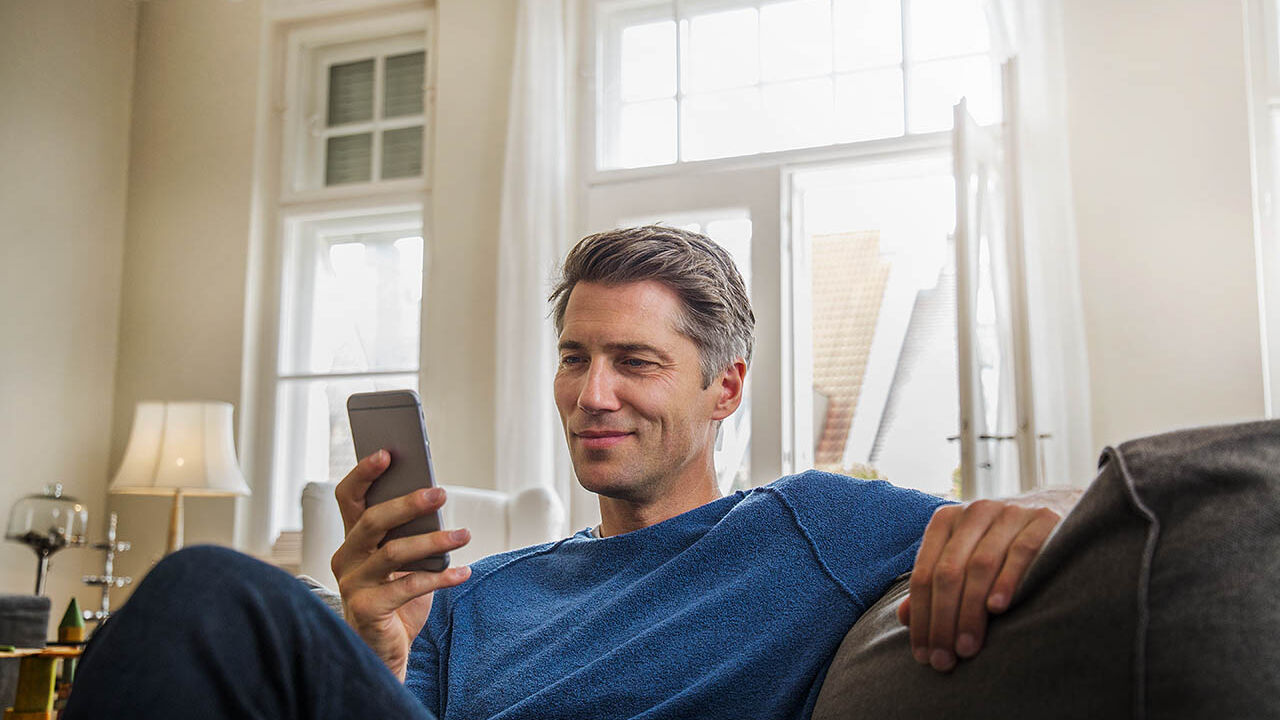 Ein Mann sitzt im Wohnzimmer auf dem Sofa und schaut in sein Handy.