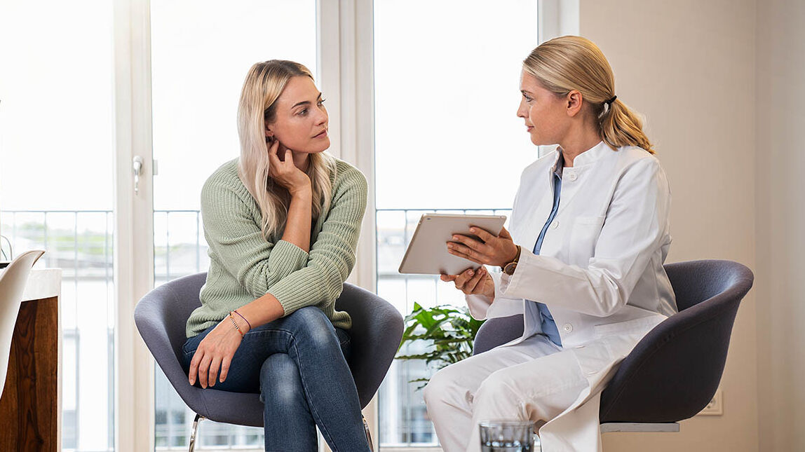 Eine Frau sitzt in einer Beratungszone der Arztpraxis und wird von ihrer Ärztin zu Vorsorgeuntersuchungen beraten.