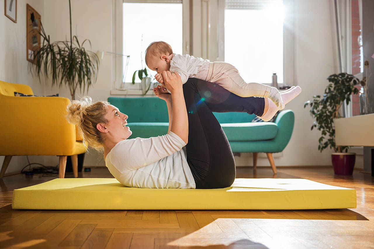 Eine Mutter macht Rückbildungsübungen mit ihrem Neugeborenen auf einer Sportmatte im Wohnzimmer.