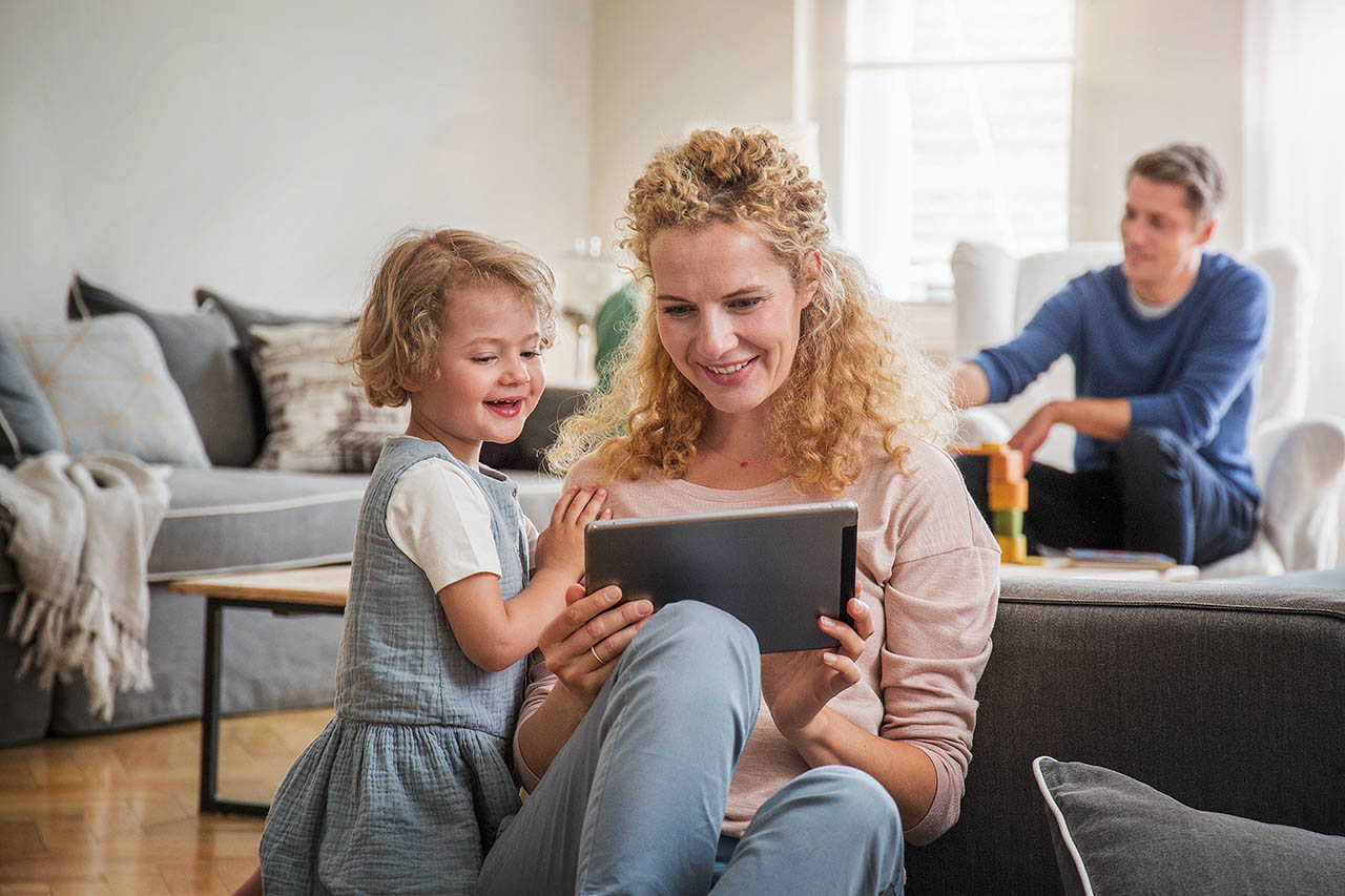 Eine Mutter sitzt mit ihrem Kleinkind auf dem Wohnzimmerboden und sie schauen gemeinsam in ein Tablet.
