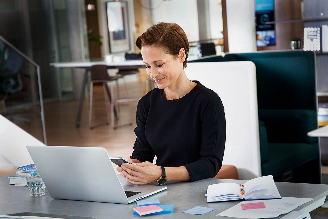 Eine Frau sitzt in einem modernen Büro vor ihrem Laptop und schaut lächelnd auf ihr Handy.