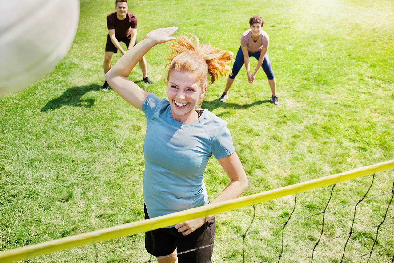Eine Frau spielt mit Freunden im Park Volleyball und springt gerade hoch, um den Ball über das Netzt zu schlagen..