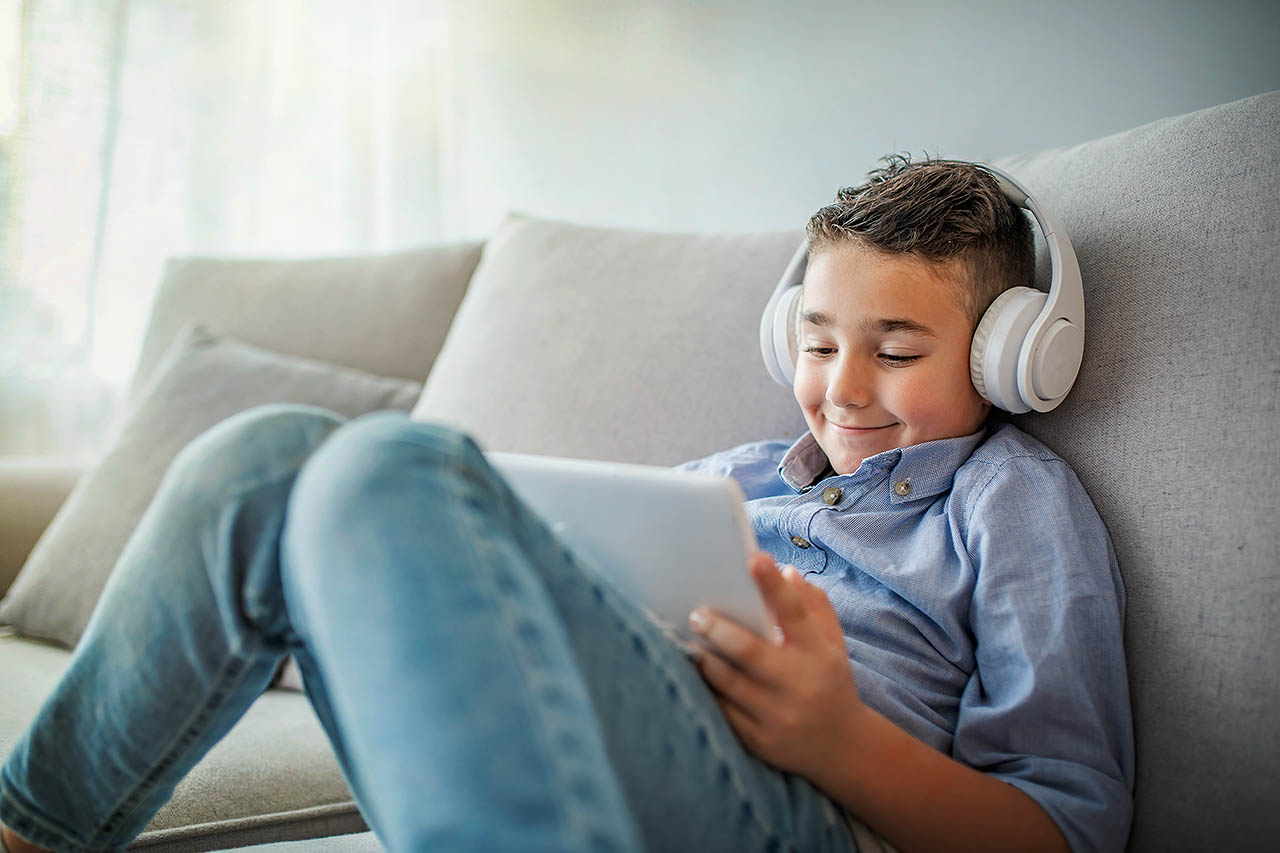 Ein Junge sitzt mit Kopfhörer und einem Tablet auf dem Sofa und lächelt.