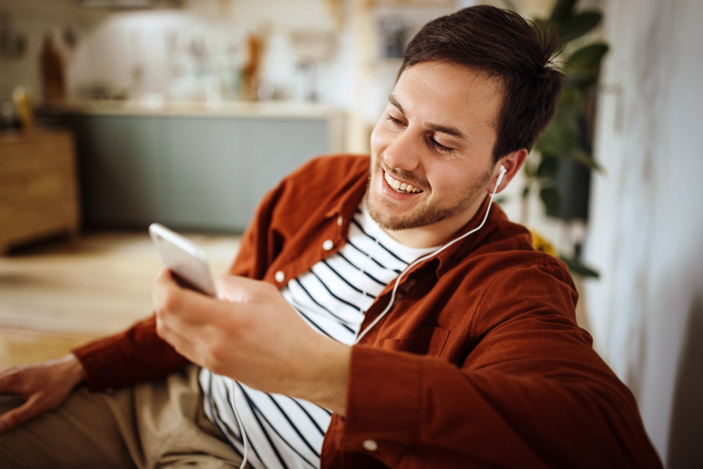 Ein Mann sitzt lachend mit seinem Mobiltelefon in auf dem Sofa und hat Kopfhörer in den Ohren.