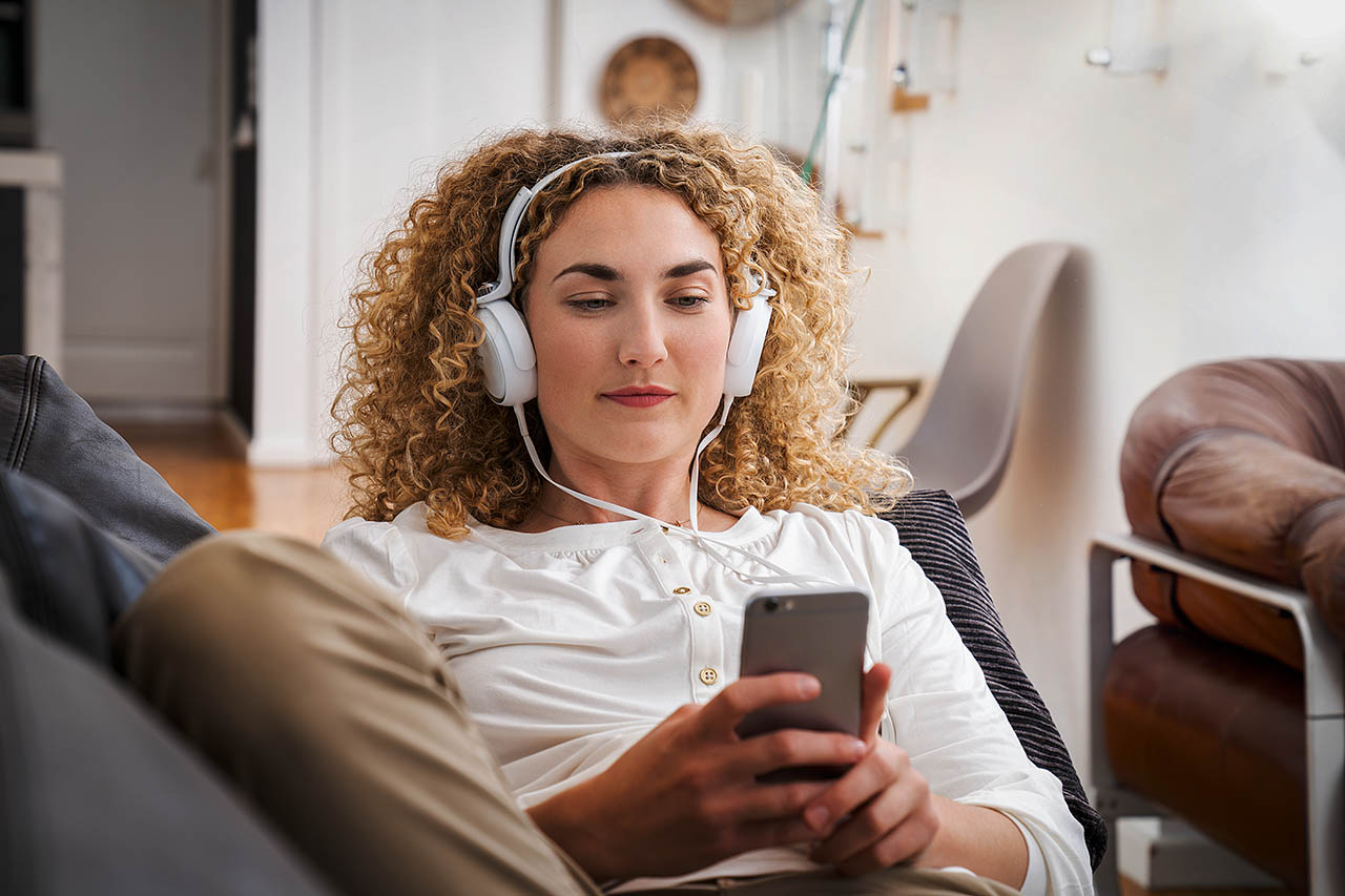 Eine Frau sitzt mit Kopfhörern auf dem Sofa und schaut auf ihr Handy.